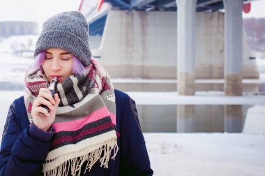 Vaping kız. Kış sokak portre bir kadın hipster, mor boyalı saçlı, gri bir şapka ve atkı ördü. kadın sokakta karla kaplı Nehri yakınında elektronik sigara içiyor