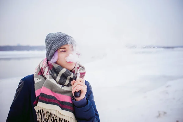 Vaping dívka. Zimní pouliční portrét ženy bederní, fialová barvené vlasy, šedé pletené čepice a šála. žena kouří elektronické cigarety v ulici poblíž řeky zasněžených — Stock fotografie