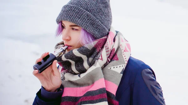 Rapariga vaporizadora. retrato de rua inverno de uma mulher hipster, cabelo tingido de roxo, um chapéu de malha cinza e cachecol. mulher fuma um cigarro eletrônico na rua perto do rio coberto de neve — Fotografia de Stock