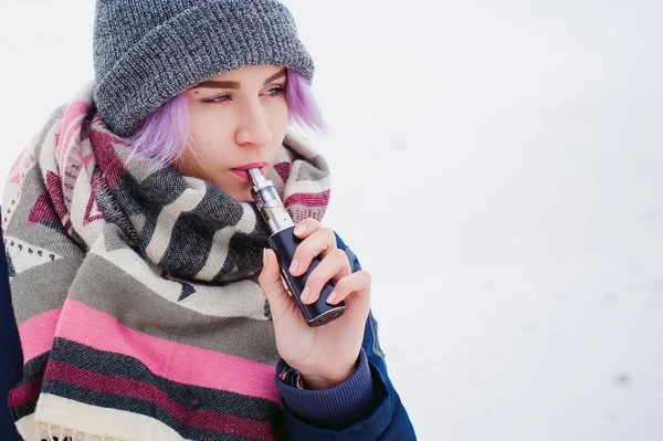 Vaping kız. Kış sokak portre bir kadın hipster, mor boyalı saçlı, gri bir şapka ve atkı ördü. kadın sokakta karla kaplı Nehri yakınında elektronik sigara içiyor — Stok fotoğraf