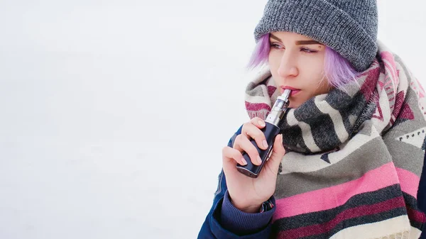 Rapariga vaporizadora. retrato de rua inverno de uma mulher hipster, cabelo tingido de roxo, um chapéu de malha cinza e cachecol. mulher fuma um cigarro eletrônico na rua perto do rio coberto de neve — Fotografia de Stock