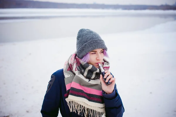 Rapariga vaporizadora. retrato de rua inverno de uma mulher hipster, cabelo tingido de roxo, um chapéu de malha cinza e cachecol. mulher fuma um cigarro eletrônico na rua perto do rio coberto de neve — Fotografia de Stock