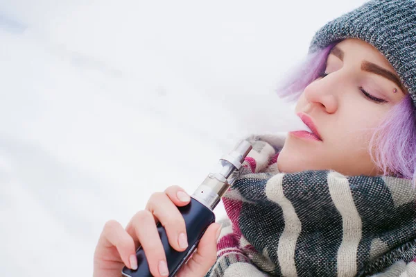 Rapariga vaporizadora. retrato de rua inverno de uma mulher hipster, cabelo tingido de roxo, um chapéu de malha cinza e cachecol. mulher fuma um cigarro eletrônico na rua perto do rio coberto de neve — Fotografia de Stock