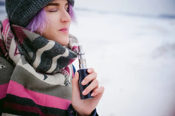 Vaping kız. Kış sokak portre bir kadın hipster, mor boyalı saçlı, gri bir şapka ve atkı ördü. kadın sokakta karla kaplı Nehri yakınında elektronik sigara içiyor — Stok fotoğraf