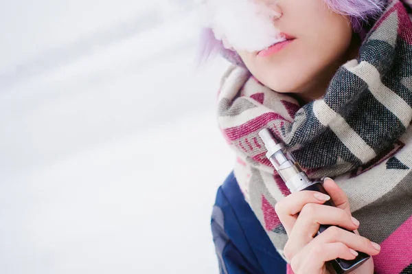 Dampfende Mädchen. Winterliches Straßenporträt einer Hipsterin, lila gefärbtes Haar, graue Strickmütze und Schal. Frau raucht auf der Straße in der Nähe des schneebedeckten Flusses eine elektronische Zigarette — Stockfoto