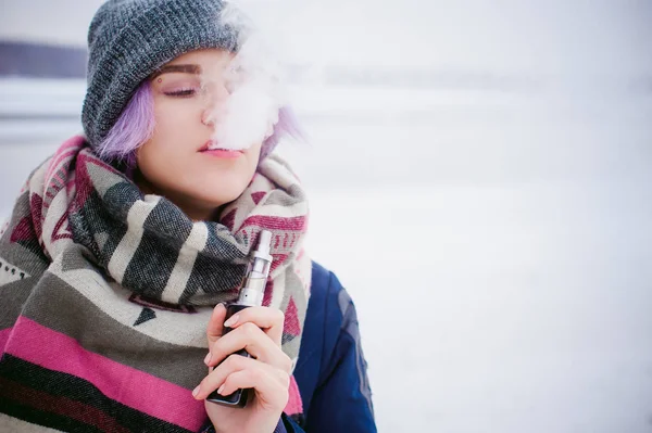 Chica de vapeo. retrato de invierno calle de una mujer hipster, pelo teñido de púrpura, un sombrero de punto gris y bufanda. mujer fuma un cigarrillo electrónico en la calle cerca del río cubierto de nieve — Foto de Stock
