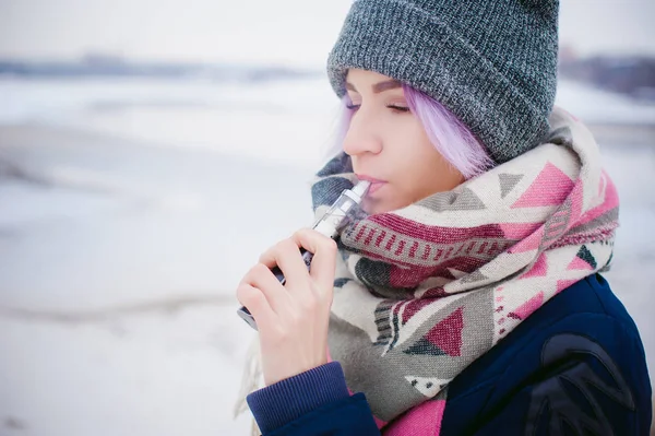 Vaping flicka. vinter gatan porträtt av en kvinna hipster, lila-färgade hår, en grå Stickad mössa och halsduk. kvinnan röker en elektronisk cigarett i gata nära floden snötäckta — Stockfoto