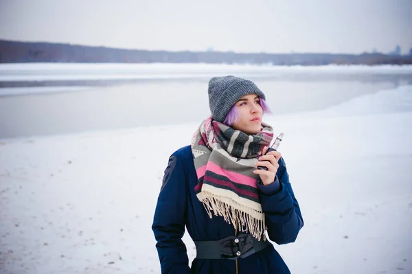Gadis penghisap. potret musim dingin seorang wanita hipster, rambut berwarna ungu, topi rajutan abu-abu dan syal. Wanita merokok rokok elektronik di jalan dekat sungai bersalju — Stok Foto