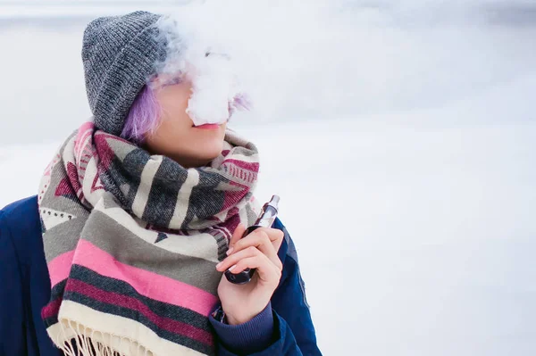 Vaping flicka. vinter gatan porträtt av en kvinna hipster, lila-färgade hår, en grå Stickad mössa och halsduk. kvinnan röker en elektronisk cigarett i gata nära floden snötäckta — Stockfoto