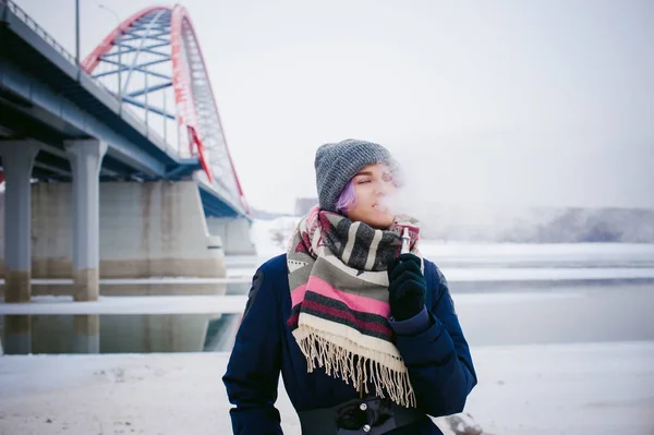 Gadis penghisap. potret musim dingin seorang wanita hipster, rambut berwarna ungu, topi rajutan abu-abu dan syal. Wanita merokok rokok elektronik di jalan dekat sungai bersalju — Stok Foto