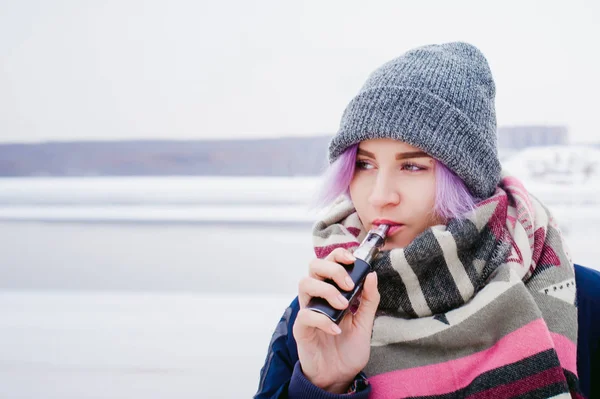 Vaping flicka. vinter gatan porträtt av en kvinna hipster, lila-färgade hår, en grå Stickad mössa och halsduk. kvinnan röker en elektronisk cigarett i gata nära floden snötäckta — Stockfoto