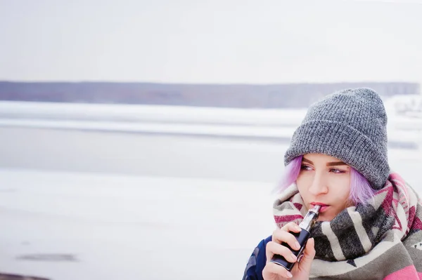 Vaping lány. tél utca portréja nő csípő, lila-festett haj, egy szürke kötött sapka és sál. nő dohányzik az utcán, a havas-folyó közelében elektronikus cigaretta — Stock Fotó