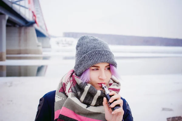Vaping flicka. vinter gatan porträtt av en kvinna hipster, lila-färgade hår, en grå Stickad mössa och halsduk. kvinnan röker en elektronisk cigarett i gata nära floden snötäckta — Stockfoto