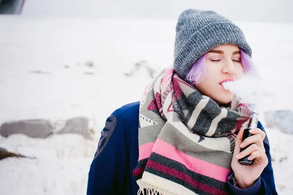 Rapariga vaporizadora. retrato de rua inverno de uma mulher hipster, cabelo tingido de roxo, um chapéu de malha cinza e cachecol. mulher fuma um cigarro eletrônico na rua perto do rio coberto de neve — Fotografia de Stock