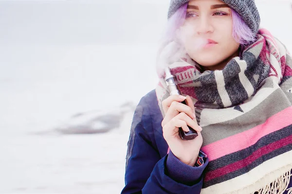 Vaping girl. portrait de rue d'hiver d'une femme hipster, cheveux teints en violet, un chapeau et une écharpe gris tricoté. femme fume une cigarette électronique dans la rue près de la rivière enneigée — Photo