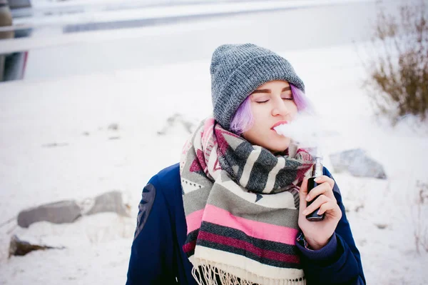 Rapariga vaporizadora. retrato de rua inverno de uma mulher hipster, cabelo tingido de roxo, um chapéu de malha cinza e cachecol. mulher fuma um cigarro eletrônico na rua perto do rio coberto de neve — Fotografia de Stock