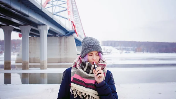 Vaping dívka. Zimní pouliční portrét ženy bederní, fialová barvené vlasy, šedé pletené čepice a šála. žena kouří elektronické cigarety v ulici poblíž řeky zasněžených — Stock fotografie