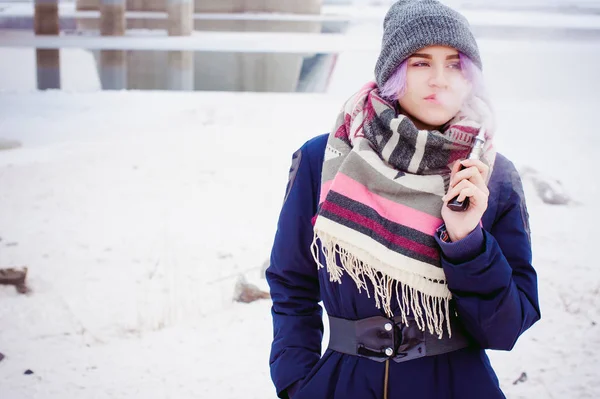 Κορίτσι ατμίσματος. χειμώνα δρόμου πορτρέτο του ένα hipster γυναίκα, μαλλιά βαμμένα μωβ, ένα γκρι πλεκτό καπέλο και κασκόλ. γυναίκα που καπνίζει ηλεκτρονικό τσιγάρο στο δρόμο κοντά στο χιονισμένο ποτάμι — Φωτογραφία Αρχείου