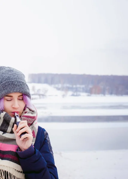 Ragazza vaporizzatrice. ritratto di strada invernale di una donna hipster, capelli tinti di viola, un cappello a maglia grigio e sciarpa. donna fuma una sigaretta elettronica nella strada vicino al fiume innevato — Foto Stock