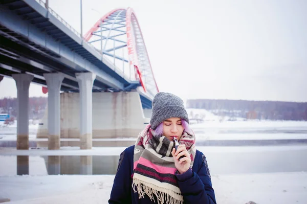 Vaping girl. portrait de rue d'hiver d'une femme hipster, cheveux teints en violet, un chapeau et une écharpe gris tricoté. femme fume une cigarette électronique dans la rue près de la rivière enneigée — Photo