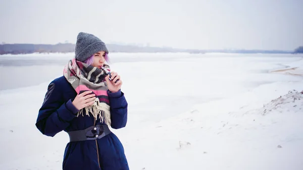 Κορίτσι ατμίσματος. χειμώνα δρόμου πορτρέτο του ένα hipster γυναίκα, μαλλιά βαμμένα μωβ, ένα γκρι πλεκτό καπέλο και κασκόλ. γυναίκα που καπνίζει ηλεκτρονικό τσιγάρο στο δρόμο κοντά στο χιονισμένο ποτάμι — Φωτογραφία Αρχείου