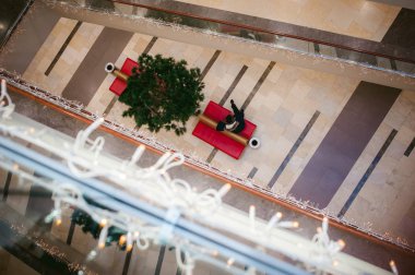 bir alışveriş merkezinde bir cep telefonu ile bir kırmızı kanepede elinde, Noel ağacının yanında oturan bir adam Noel ışıkları ile dekore edilmiştir. yukarıdan görüntülemek