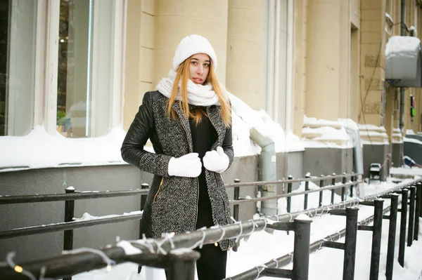 Portrait of a cute woman in white scarf and hat knitted coat on the street on a background buildings — Stock Photo, Image