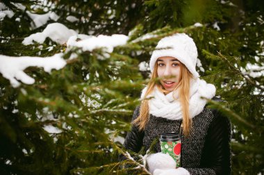 Beyaz atkı ve şapka sevimli kadın portresi kat kar açık havada arka plan üzerinde örme ve kışın çam dalları bulanık. Kız elinde, Noel paket servisi olan restoran kahve bir fincan kahve tutar