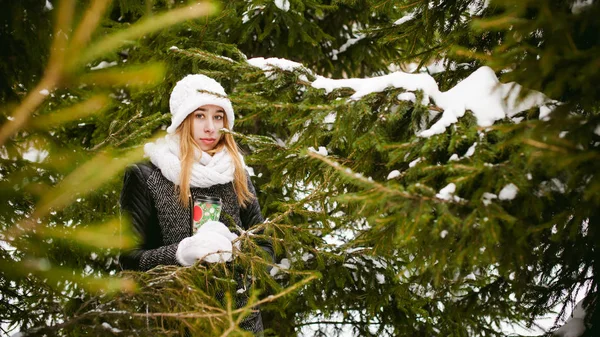 Portrait of cute woman in white scarf and hat knitted coat on outdoors background of snow and blurred fir branches in winter. — Stock Photo, Image