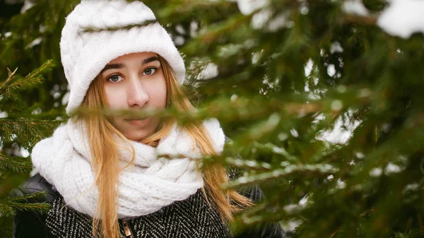 Ritratto di donna carina in sciarpa bianca e cappello cappotto lavorato a maglia su sfondo esterno di neve e rami di abete sfocati in inverno . — Foto Stock