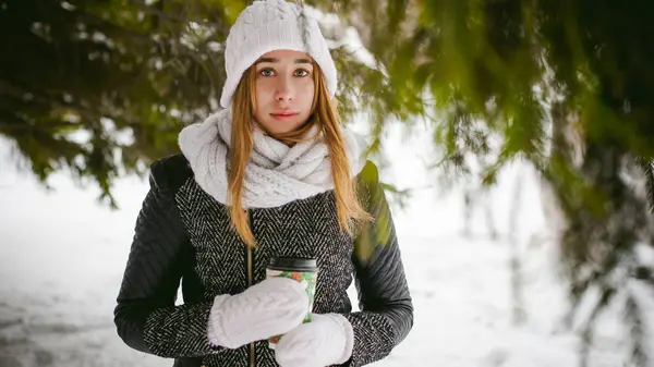 白いスカーフと帽子でかわいい女性の肖像ニット雪の屋外背景にコートと冬のモミの枝をぼやけています。女の子がクリスマスのテイクアウト コーヒーの手でコーヒー カップを保持しています。 — ストック写真