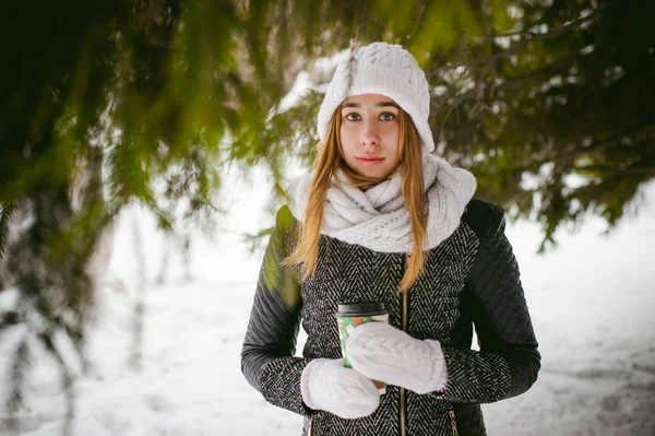 Portrait de jolie femme en écharpe blanche et chapeau manteau tricoté sur fond extérieur de neige et branches de sapin floues en hiver. fille tient une tasse de café dans la main, café à emporter de Noël — Photo