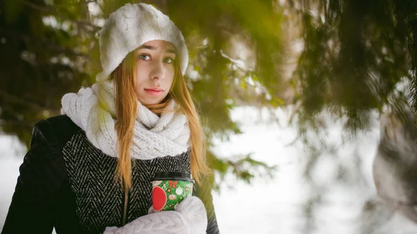 Портрет милой женщины в белом шарфе и шапке вязаном пальто на открытом воздухе на фоне снега и размытых еловых ветвей зимой . — стоковое фото