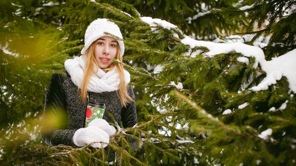 Porträtt av söt kvinna i vit halsduk och mössa stickad kappa på utomhus bakgrund av snö och suddig fir grenar på vintern — Stockfoto