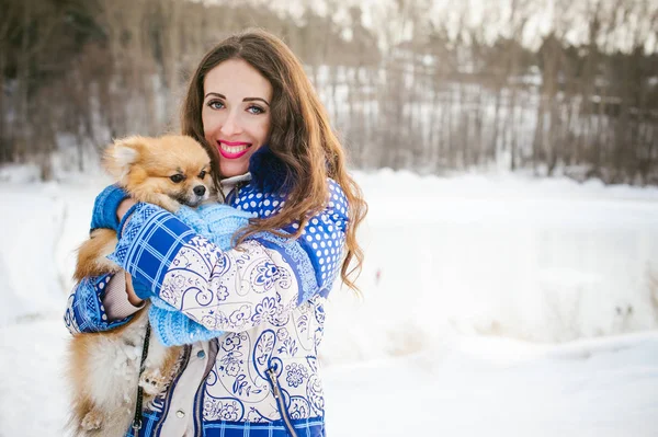 smiling woman holding her pet dog in his hands near face, hugging. Spitz breed dog playing with a woman walking outdoors winter day, warm clothing. love and care for the pet, dog walking