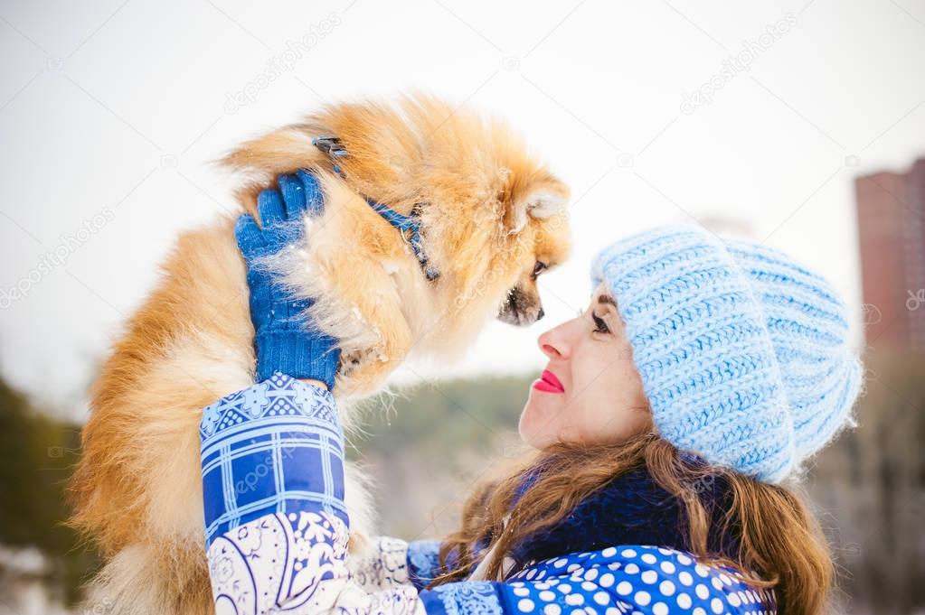 smiling woman holding her pet dog in his hands near face, overhead. Spitz breed dog playing with a woman walking outdoors winter day, warm clothing. love and care for the pet, dog walking