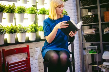 sarışın kadın elbiseli bir sandalye bir kitapla elinde, houseplants fonunda oturur. okuma meşgul