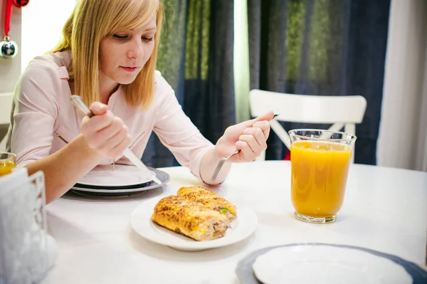 Pretty blonde woman in a pink shirt, eating at home a fork and knife. breakfast sandwich and peach juice. on white table in bright interior of kitchen in plate is sandwich juice poured in glass — Stock Photo, Image