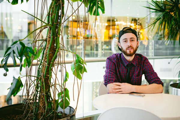 bir ekose gömlek ve kravat ve palmiye ağaçları arka plan üzerinde alışveriş balkonunda bir kafede bir masada oturuyor kap, sakallı genç adam