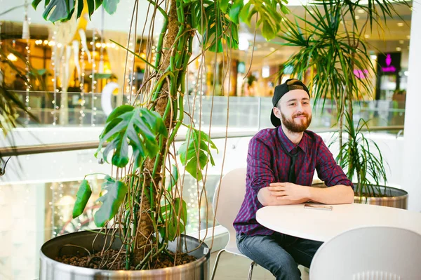 bir ekose gömlek ve kravat ve palmiye ağaçları arka plan üzerinde alışveriş balkonunda bir kafede bir masada oturuyor kap, sakallı genç adam