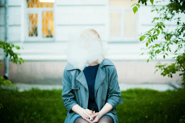 Ritratto all'aperto di giovane donna bionda, indossa un impermeabile, fuma sigaretta elettronica — Foto Stock