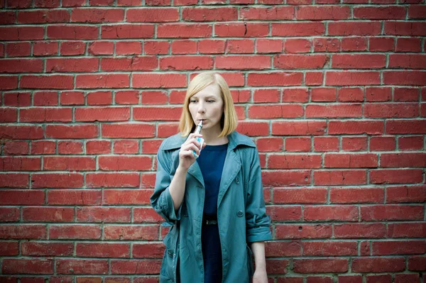 Retrato ao ar livre de jovem loira, vestindo uma capa de chuva, fumar cigarro eletrônico — Fotografia de Stock