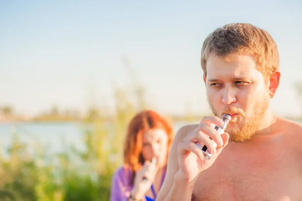 Pareja joven descansando en la playa, fumando cigarrillo electrónico — Foto de Stock