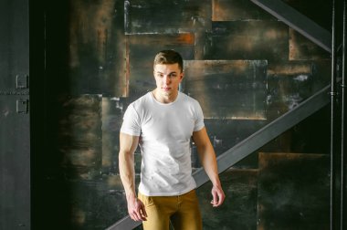 Genç seksi erkek vücut geliştirmeci atlet, stilize bir metal duvar arka plan üzerinde bir çatı katında stüdyo portre, bir adam model beyaz bir T-shirt ve kahverengi pantolon