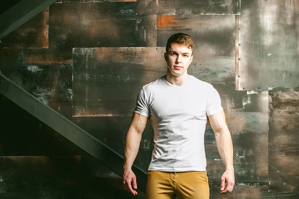 Young sexy men bodybuilder athlete, Studio portrait in a loft on the background of a stylized metal wall, a guy model in a white T-shirt and brown trousers — Stock Photo, Image