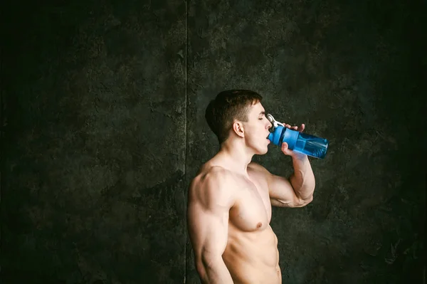 年轻的性感男士健美运动员，工作室肖像阁楼上的程式化的墙，家伙模型与裸露躯干饮料水从运动水壶训练后的蓝颜色的背景 — 图库照片