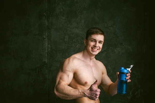 Young sexy men bodybuilder athlete,studio portrait loft on background of stylized wall,guy model With bare torso Drinks water from sports bottle of blue color after training