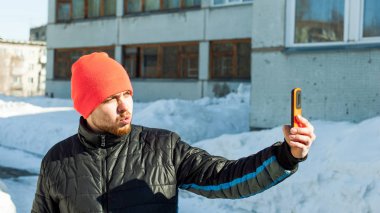Adam kendini fotoğraf çekme. bir sonbahar spor rahat ceket ve parlak turuncu şapka giymiş sakallı adam selfie telefon şık durumunda, açık havada güneşli kış gününde yürüyüş yapar