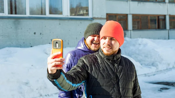 Two men photographing themselves.guy with beard,dressed in autumn sports casual jacket and bright orange hat, makes selfie on phone in stylish case,walking with his friend outdoors sunny winter day