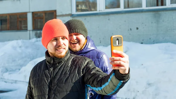 Dos hombres fotografiando themselves.guy con barba, vestido con chaqueta casual deportes de otoño y sombrero naranja brillante, hace selfie en el teléfono en caso elegante, caminando con su amigo al aire libre día de invierno soleado — Foto de Stock